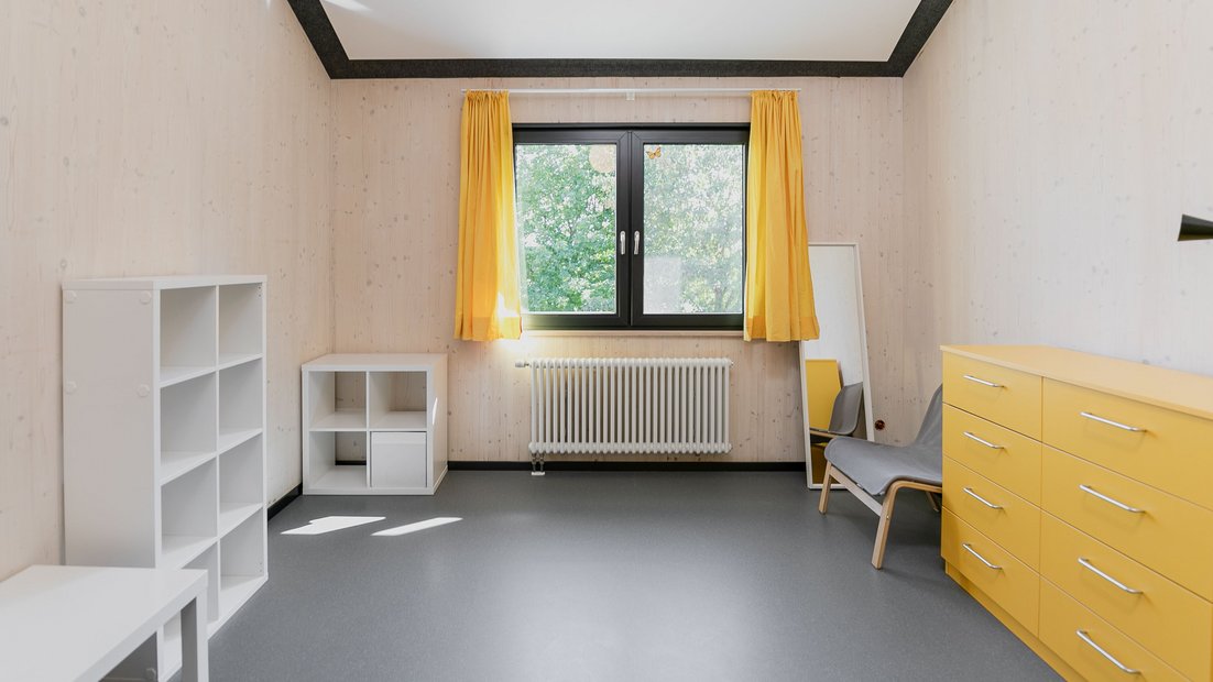 Phòng trong Rosenhaus trên lầu với tủ ngăn kéo màu vàng, kệ trắng, bàn, gương và ghế bành màu xám